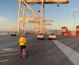 Person in a safety vest walking with luggage near cranes at a shipping port during sunset.