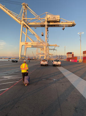 Person in a safety vest walking with luggage near cranes at a shipping port during sunset.