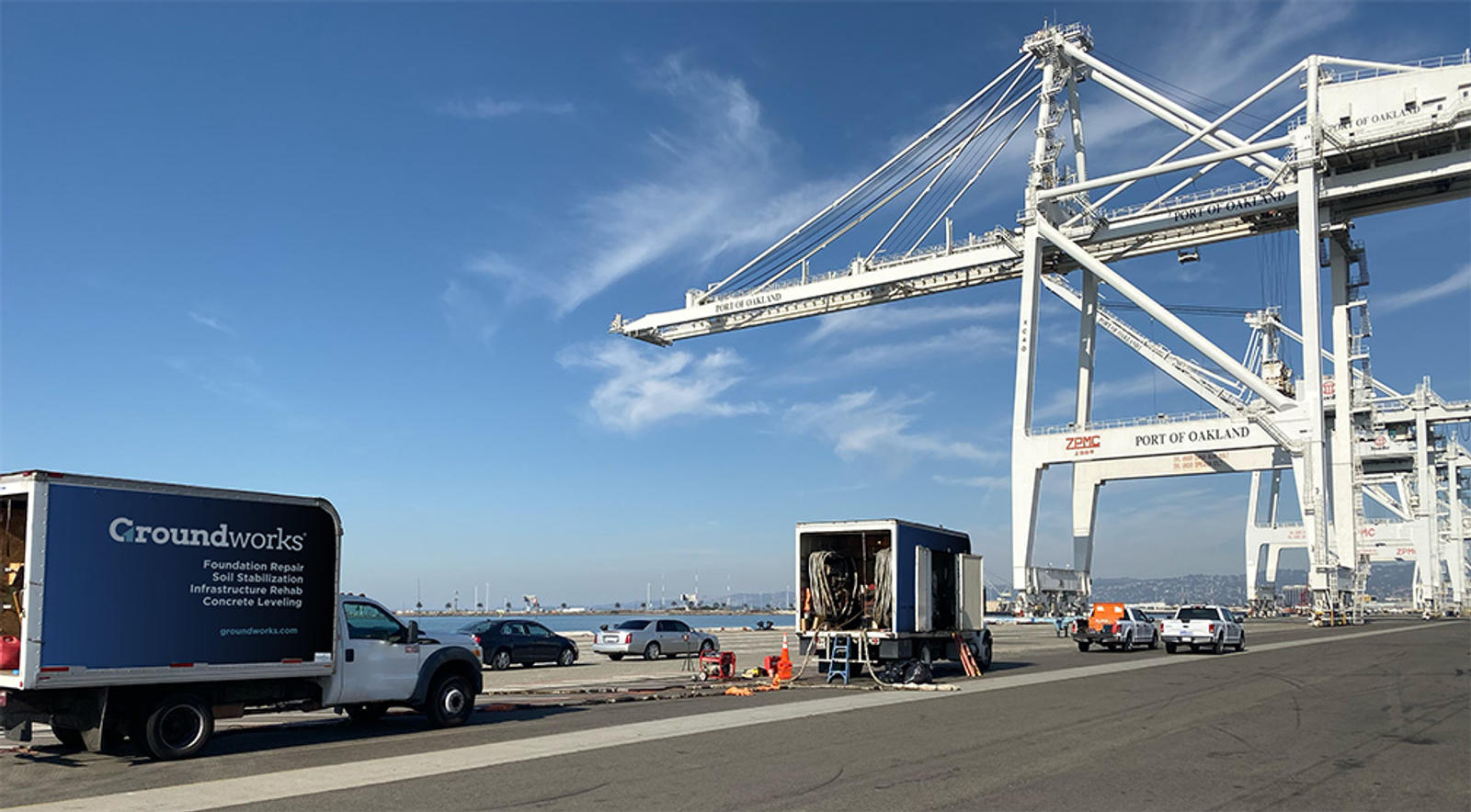 Trucks and equipment at a port with large cranes and clear blue sky in the background.
