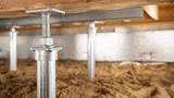 Metal support jacks reinforce a wooden floor above a dirt crawl space.