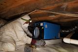Person in protective gear installs a blue dehumidifier in a crawl space with wood beams and pipes overhead.