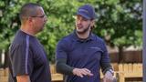 Two men talk outdoors; one gestures while explaining something to the other near a wooden fence and trees.