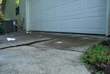 Cracked and uneven concrete driveway in front of a white garage door next to a green lawn.