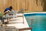 A worker repairs the edge of a backyard swimming pool using metal tools and equipment.