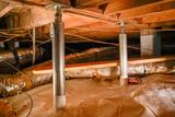 Metal support jacks reinforce wooden beams in a crawl space lined with a plastic moisture barrier.
