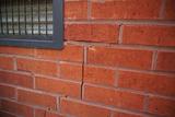 Close-up of a brick wall with visible cracks near a small window with a grid-patterned metal grate.