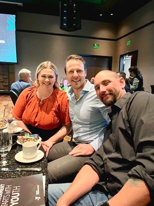 Three people smiling while seated at a table in a dimly lit room during an event.