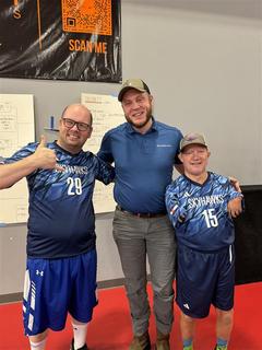 Three men smiling, two in Skyhawks sports jerseys, stand together on an indoor court.