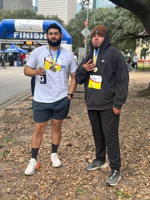 Two men with medals pose near a race finish line, one in a t-shirt and shorts, the other in a hoodie and sweatpants.