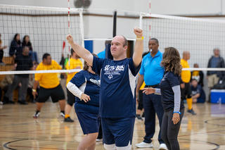 Person in a blue sports outfit celebrates on an indoor volleyball court. Other players and referees are around.