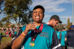 Smiling man holds a medal at an outdoor event, wearing a "Mighty Menifee" shirt. Crowd and trees in the background.