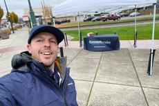 A person in a blue jacket and cap takes a selfie in front of a Groundworks tent on a cloudy day.