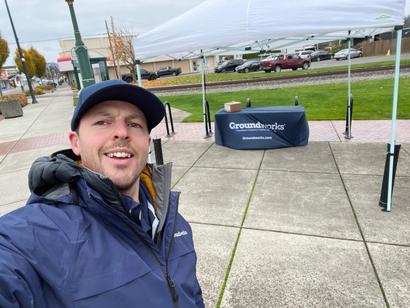A person in a blue jacket and cap takes a selfie in front of a Groundworks tent on a cloudy day.