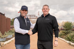 Uretek and Groundworks leaders in branded vests shake hands outdoors on a path lined with plants.