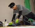 A person in work attire uses a tool on the floor while kneeling, wearing green gloves in an indoor setting.
