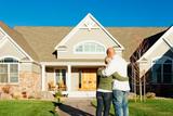 A couple stands hugging in front of a modern suburban house on a sunny day.