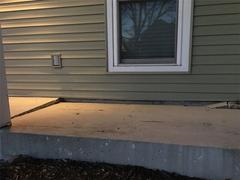 A concrete porch has a large crack running parallel to a house with green siding and a window.