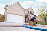 Worker in uniform repairs concrete driveway cracks outside a suburban house with an American flag.