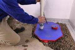 Person in blue shirt installing or inspecting a sump pump cover in a basement with gravel floor.