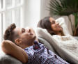 Two people relaxing on couches, hands behind their heads, near a window with a plant—a serene setting perfect for sharing basement air quality tips.