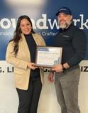 Two people smiling, holding a framed community partner certificate, standing in front of a blue sign.