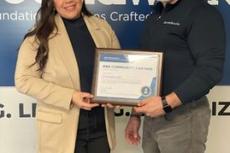 Two people smiling, holding a framed community partner certificate, standing in front of a blue sign.