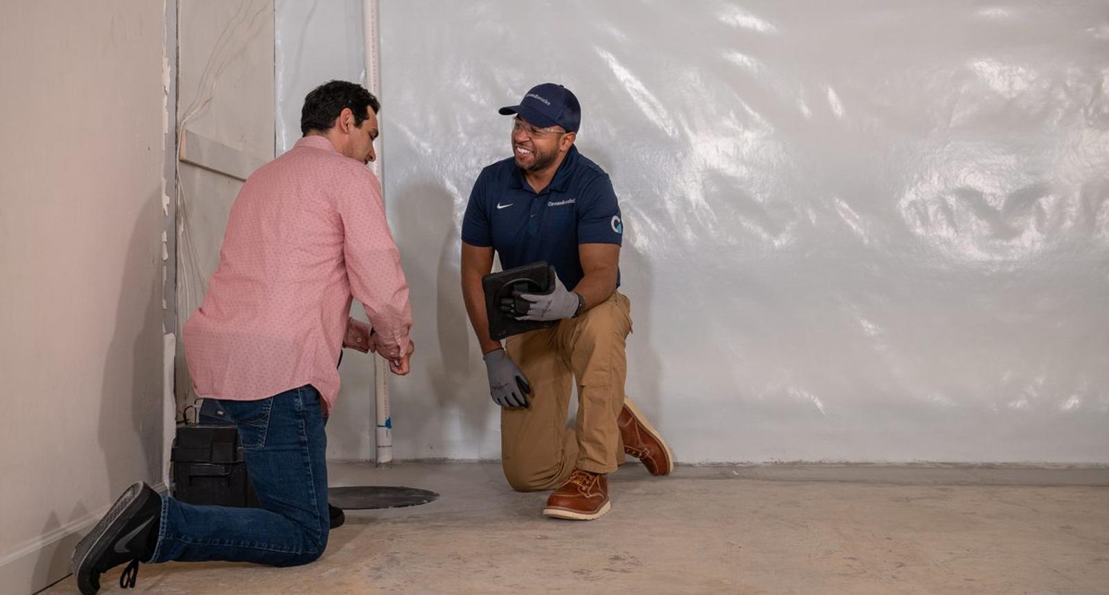 Two men kneel by a sump pump in a basement. One wears a blue uniform holding a tool, the other wears a pink shirt.