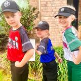 Three kids in baseball uniforms and caps, standing side by side and smiling in front of a brick building.