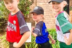 Three kids in baseball uniforms and caps, standing side by side and smiling in front of a brick building.