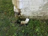 A white downspout curves by a weathered brick wall, hinting at old home foundation repair amid grass and weeds.