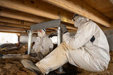 Two workers in protective suits install support beams under a wooden floor in a crawl space.