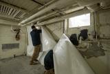 Two workers install a wall vapor barrier for foundation waterproofing in a basement with exposed pipes and a small window.