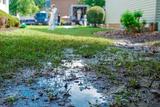 A muddy patch of grass with standing water in a yard, workers and equipment are visible in the background.