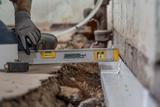Person using a level tool to check drainage alignment along a damaged concrete floor for fieldstone foundation waterproofing.
