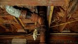 Rusty, corroded pipes and wooden beams with wood rot and signs of dry rot fungus in a crawl space.