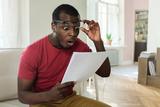 A surprised man in glasses reads a document while sitting on a bright living room couch.