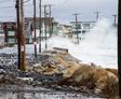 Large waves from a nor’easter crash over a seawall, causing basement flooding along a coastal street lined with houses and utility poles.