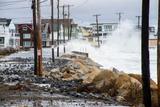 Large waves from a nor’easter crash over a seawall, causing basement flooding along a coastal street lined with houses and utility poles.