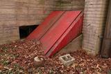 Red cellar doors surrounded by fallen leaves, beside a brick wall and cinder block—ideal for bilco door repair needs.