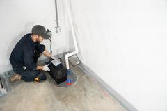 Man in work clothes inspecting a sump pump system near the concrete foundation repair area in a basement corner with white walls.