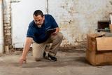 A man kneels on the ground indoors, inspecting the floor for water damage with a notepad near a cardboard box.