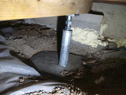 Metal support jack on a concrete footing under a wooden beam in a crawl space with exposed dirt and insulation.