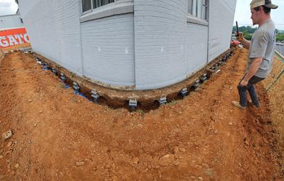A worker inspects a building foundation with exposed soil and support brackets along the base.