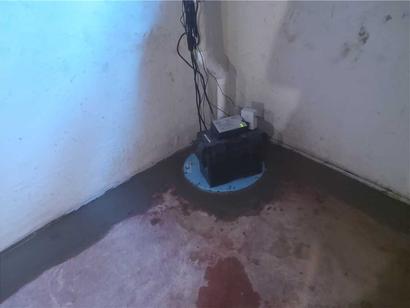 A sump pump with wires sits in a basement corner on a concrete floor near a white wall.