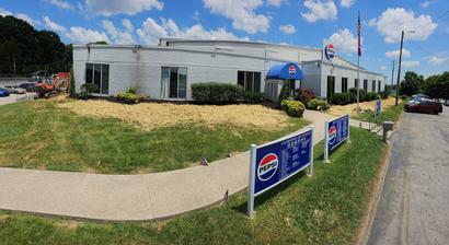 A white brick Pepsi building with blue signs, an awning, and a landscaped sidewalk under a sunny sky.