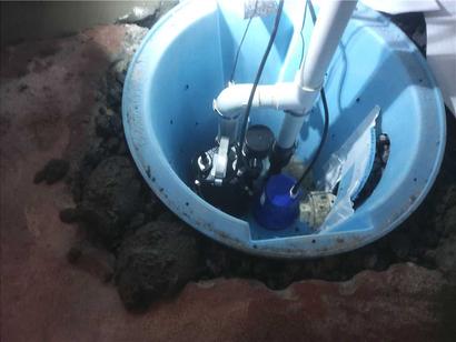 A sump pump installed in a basement pit surrounded by exposed concrete and gravel.