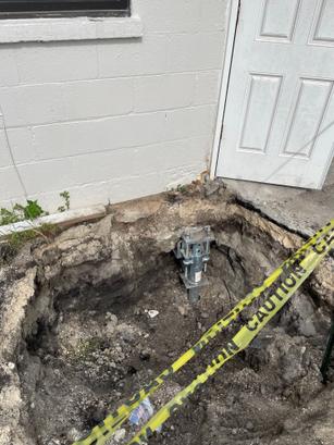 Large hole in concrete near a building, with caution tape and exposed metal footing inside the hole.