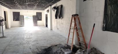Interior of a building under renovation with a ladder, broom, exposed ceiling, and plastic-covered windows.