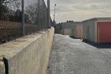 A paved alley by a chain-link fence and storage units with pink doors on a cloudy day.