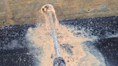 Brownish water flowing out of a pipe onto a dark, gritty surface near a concrete wall.
